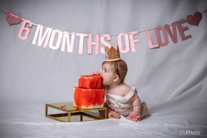 Baby in crown looks at red celebration cake under pink banner.