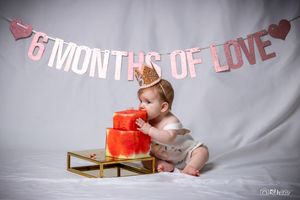 Baby reaches for red tiered cake while wearing crown under pink banner.