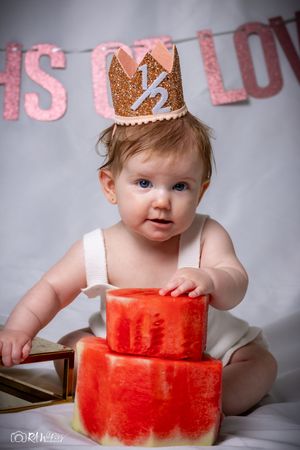 Baby in white outfit and gold crown smiles while touching red watermelon blocks against grey backdrop.