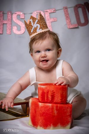 Baby in white outfit and crown poses with red tiered cake against gray backdrop.