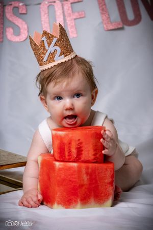 A baby wearing a sparkly gold crown poses with stacked watermelon blocks during a half birthday photoshoot.