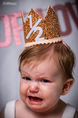 Baby wearing a gold crown makes crying face during a photo session with pink lettering backdrop.