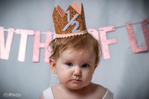 Close up of baby wearing a glittery gold crown with letters against a pink and grey background.