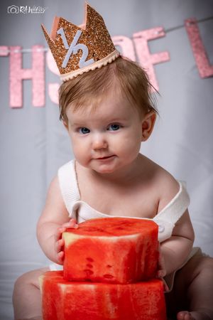 Baby in white outfit smiles while posing with watermelon blocks and wearing sparkly crown.