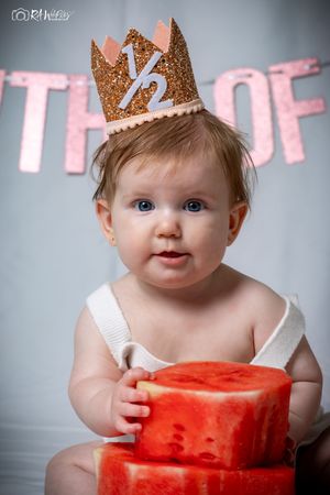 Smiling baby in white top poses with stacked watermelon props wearing a glittery crown.