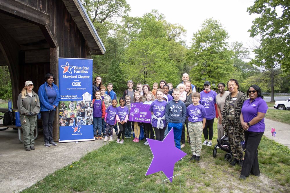 Group of adults and children in purple shirts posing outdoors near a Blue Star Families Maryland Chapter and CSX banner.
