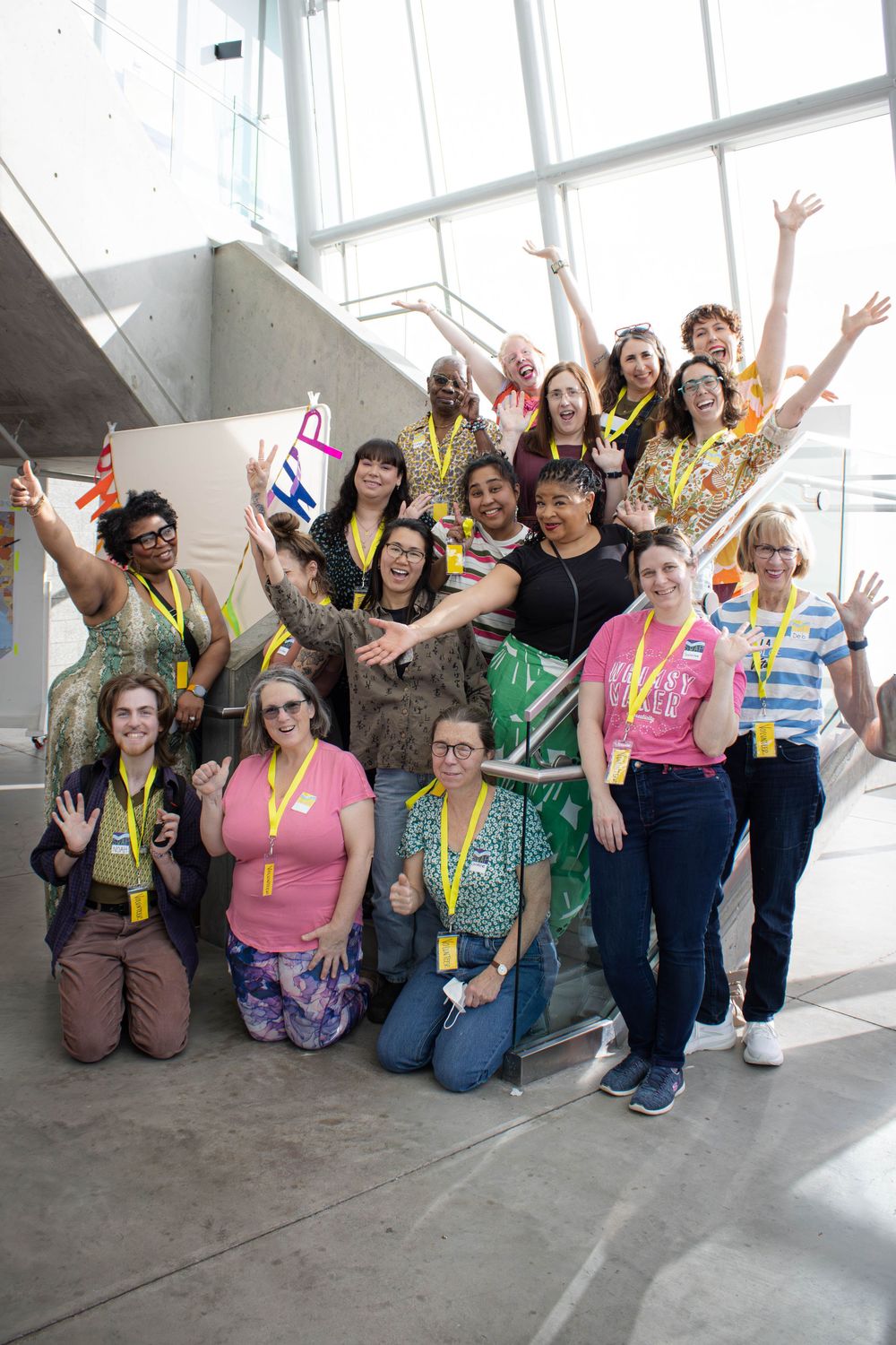 Diverse group of smiling women wearing yellow lanyards posing on stairs with arms raised in celebration.