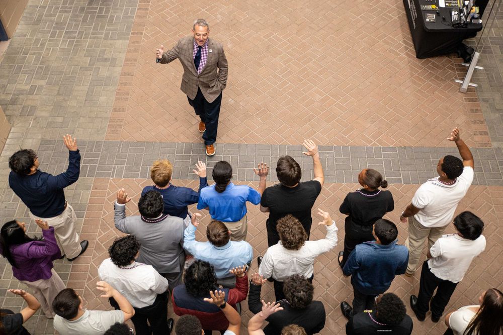 Aerial view of a speaker addressing a diverse group of people with raised hands in a brick courtyard.
