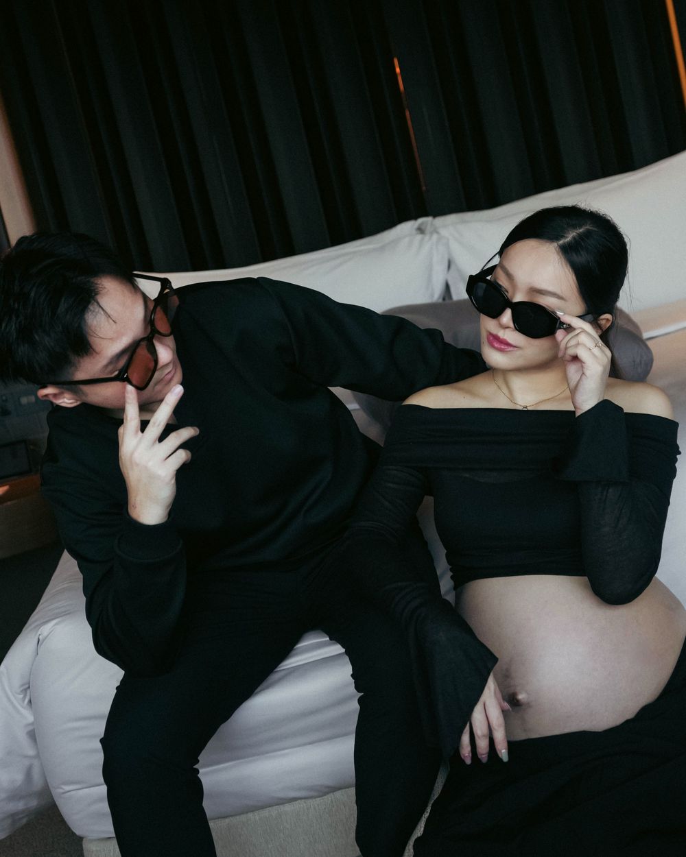 Two people wearing all black clothing and sunglasses pose together in moody, artistic photo on white couch.