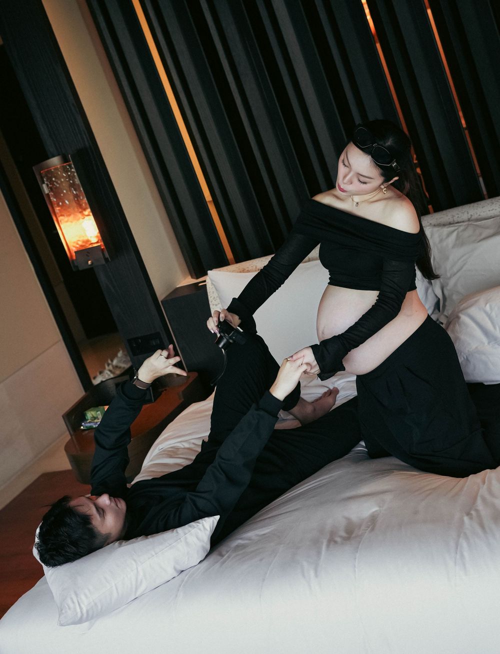 A playful maternity photoshoot in black attire on a white bed with warm ambient lighting.