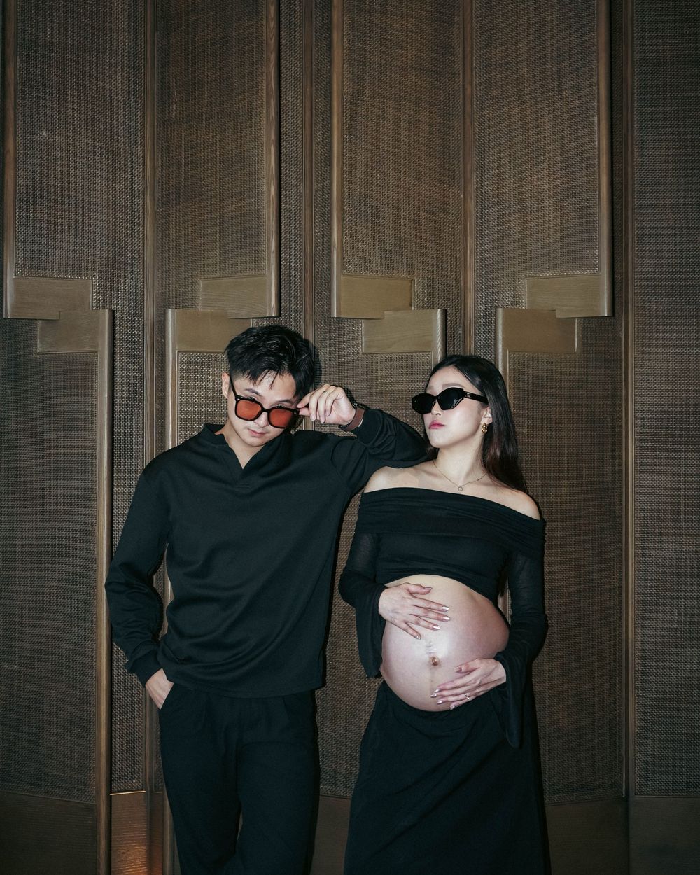 Couple in matching black outfits and sunglasses pose against wood paneled wall, showing off pregnant belly.