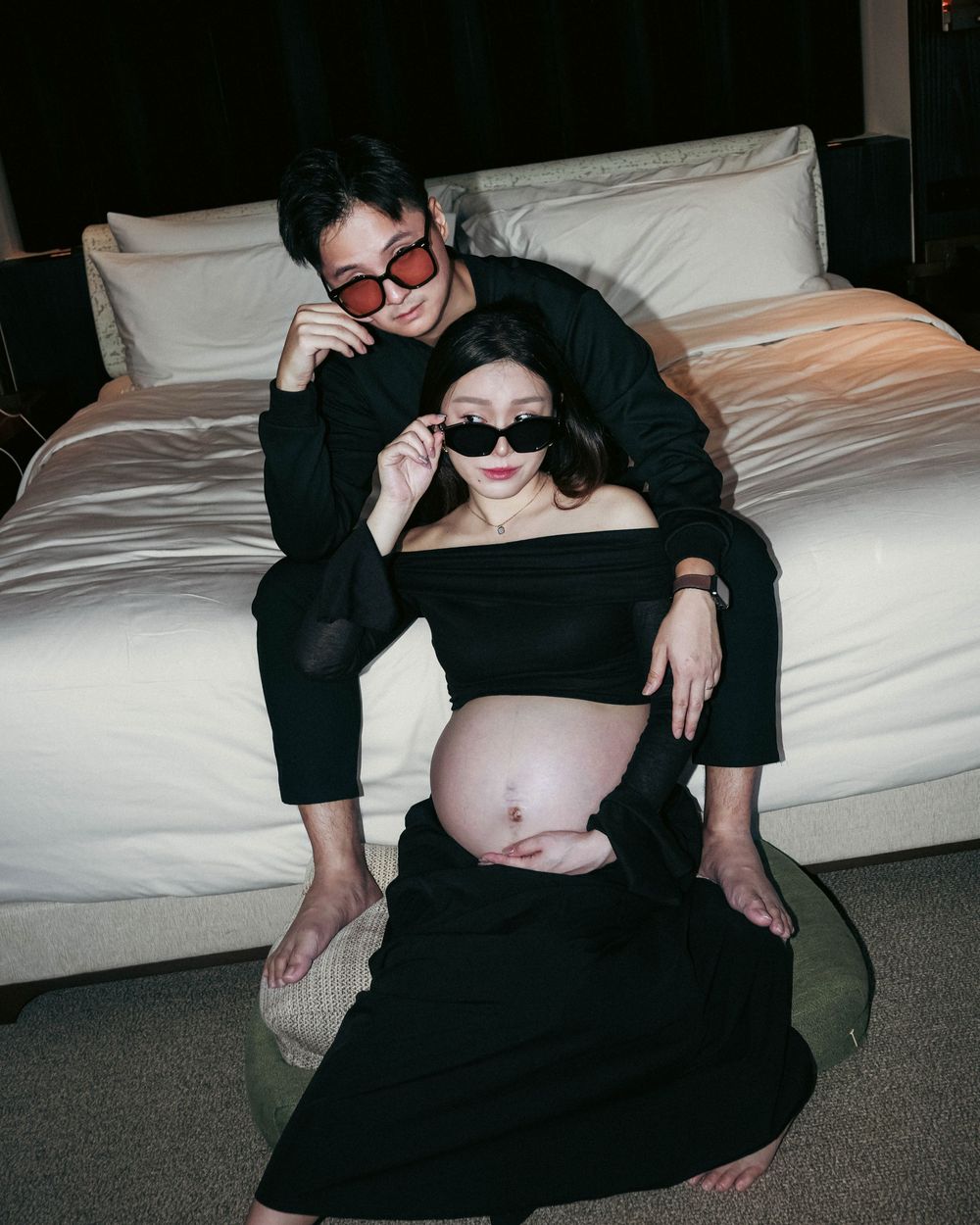 A couple wearing sunglasses and black clothing pose for a stylish maternity photo on a couch.