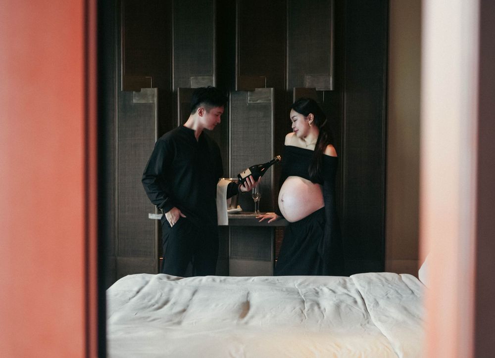 Couple stands in bedroom while expecting mother wearing black dress cradles her pregnant belly by window light.
