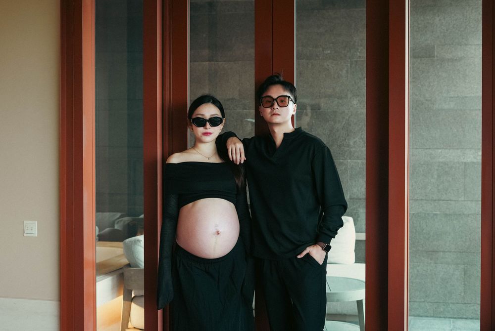 A couple in black clothing poses by a doorway with one showing a pregnant belly and both wearing sunglasses.