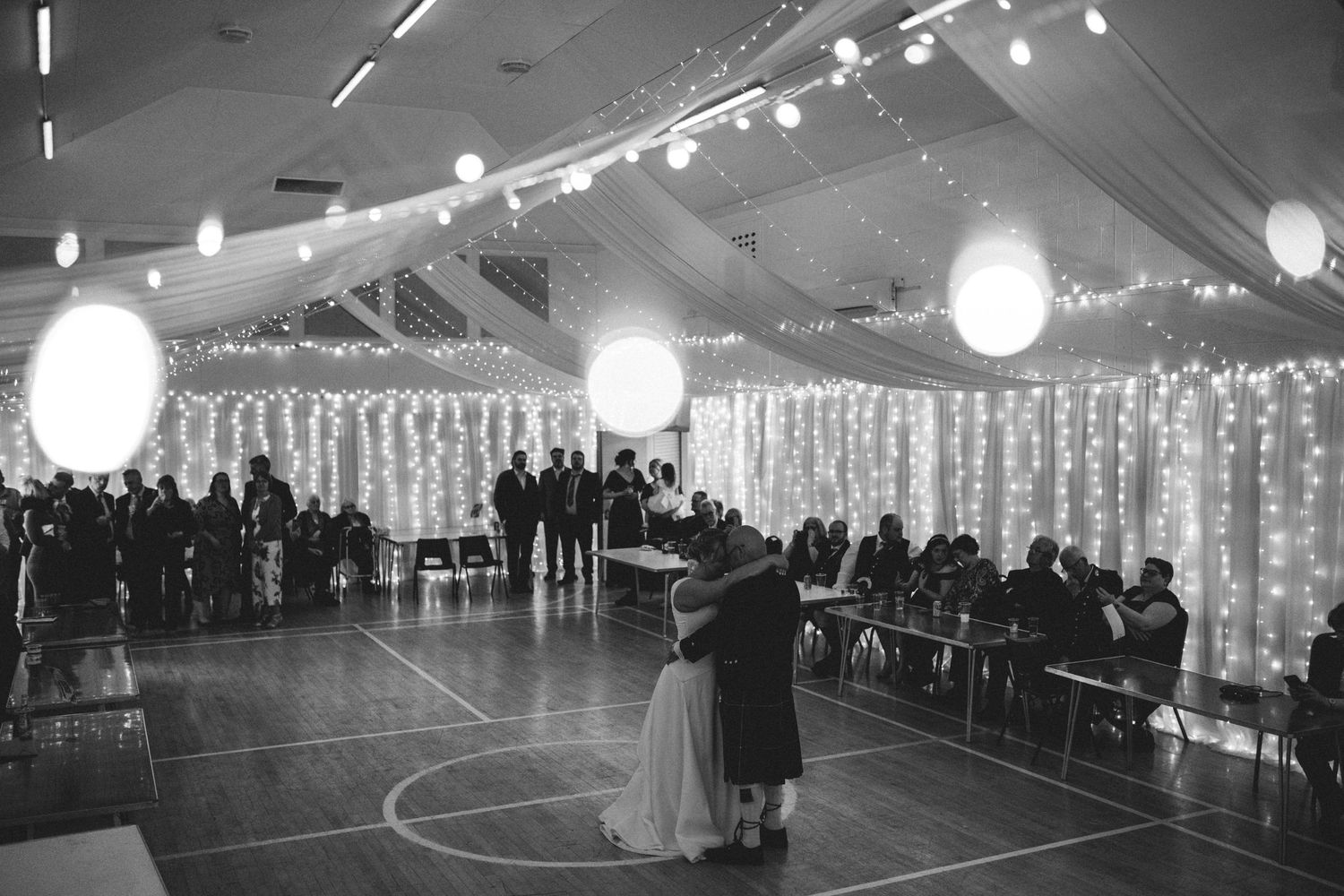 A couple dances alone on a basketball court decorated with string lights and paper lanterns for an elegant reception.