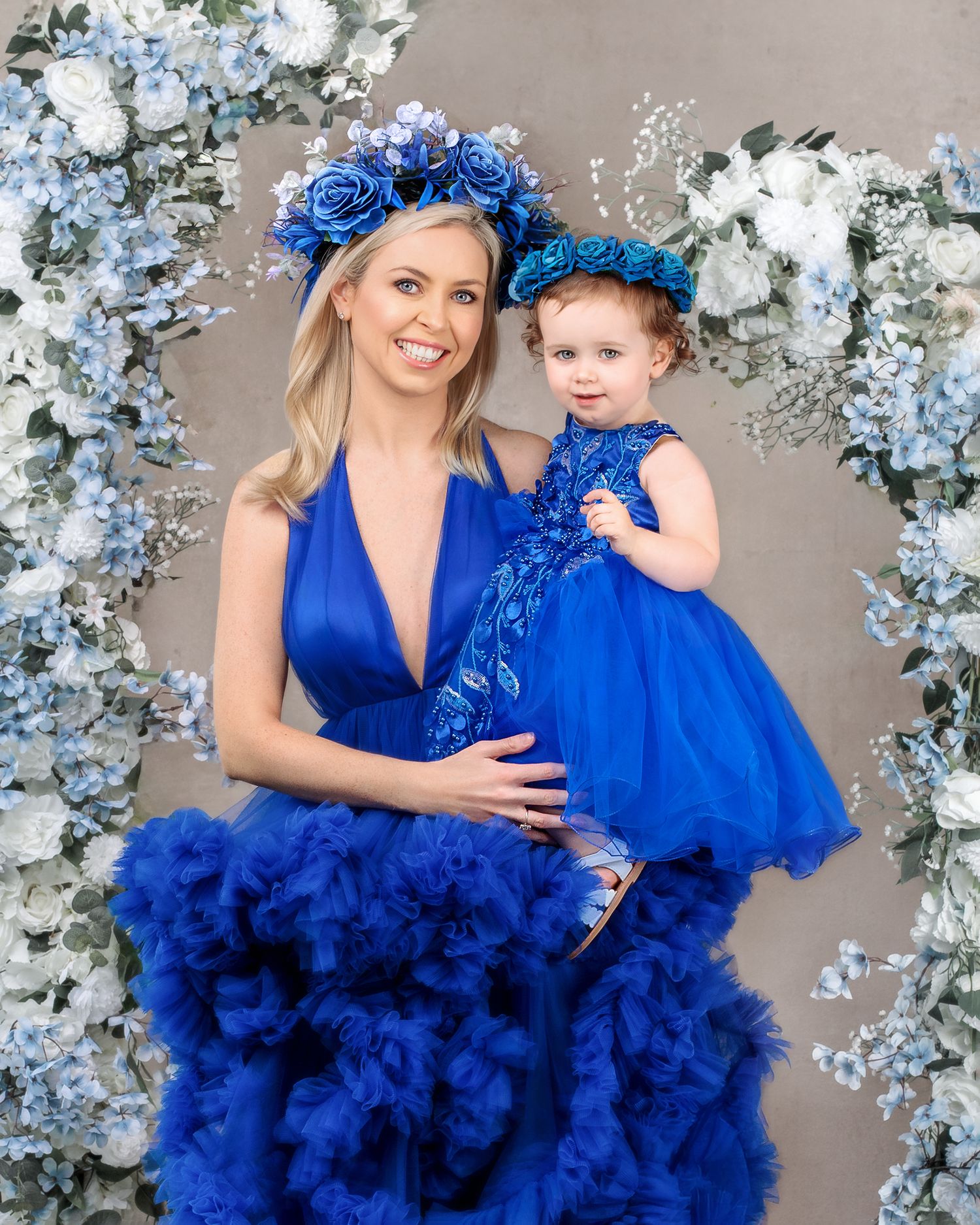 Fine art portrait: mother and child's faces close together, matching flower crowns and gowns, studio shot.