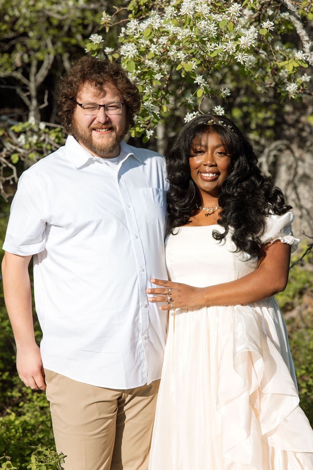 A couple poses together under blooming white spring blossoms in an outdoor garden setting.