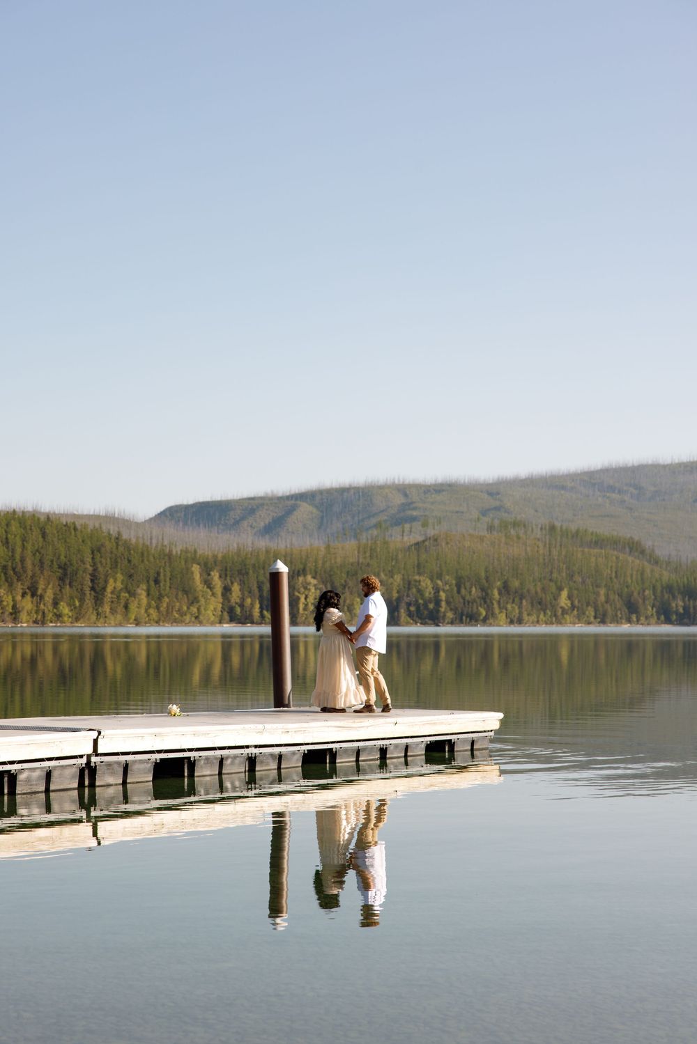 Two figures standing on a wooden dock over a serene mountain lake with forest and hills in the background.