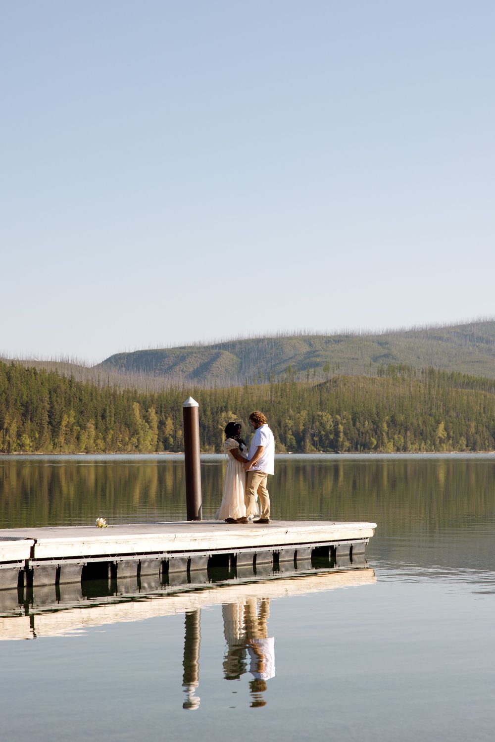 Couple stands together on a wooden dock overlooking a serene mountain lake with forest and hills in the background.