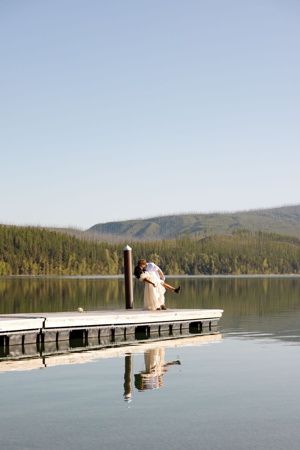 A couple in wedding attire embraces on a wooden dock overlooking a serene mountain lake during sunset.