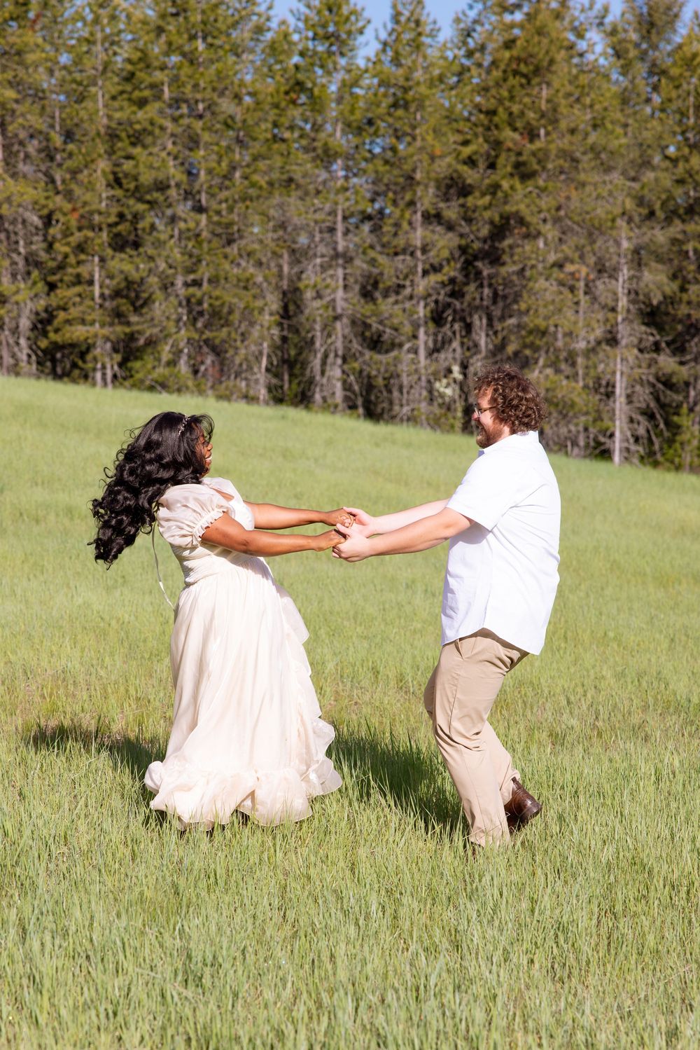 A couple holds hands while spinning together playfully in a grassy meadow with evergreen trees in the background.