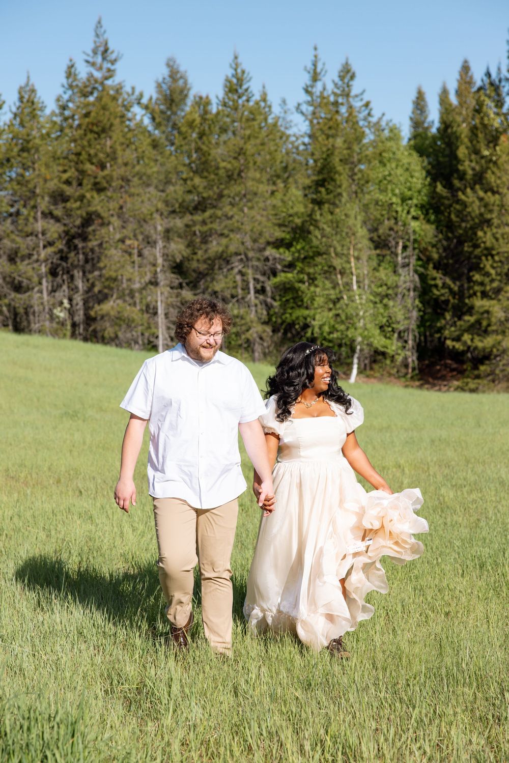 A couple walks together through a green meadow with evergreen trees in the background on a sunny day.