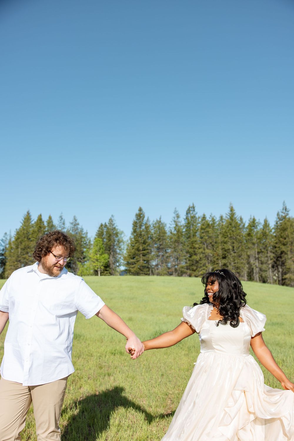 A couple holds hands while standing in a green meadow with evergreen trees and blue sky in the background.
