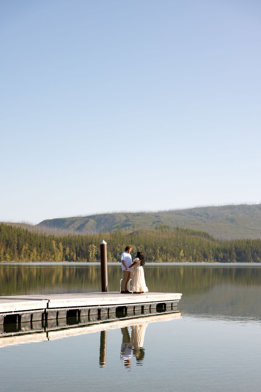 A couple stands together on a wooden dock overlooking a serene mountain lake with their reflection visible in the calm water.