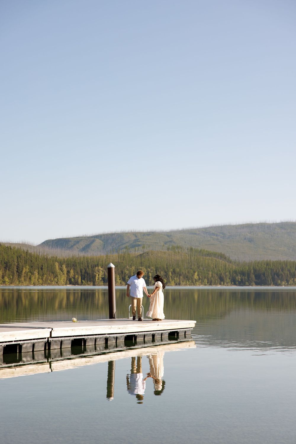 Couple standing on wooden dock overlooking serene mountain lake with perfect reflection in calm waters.