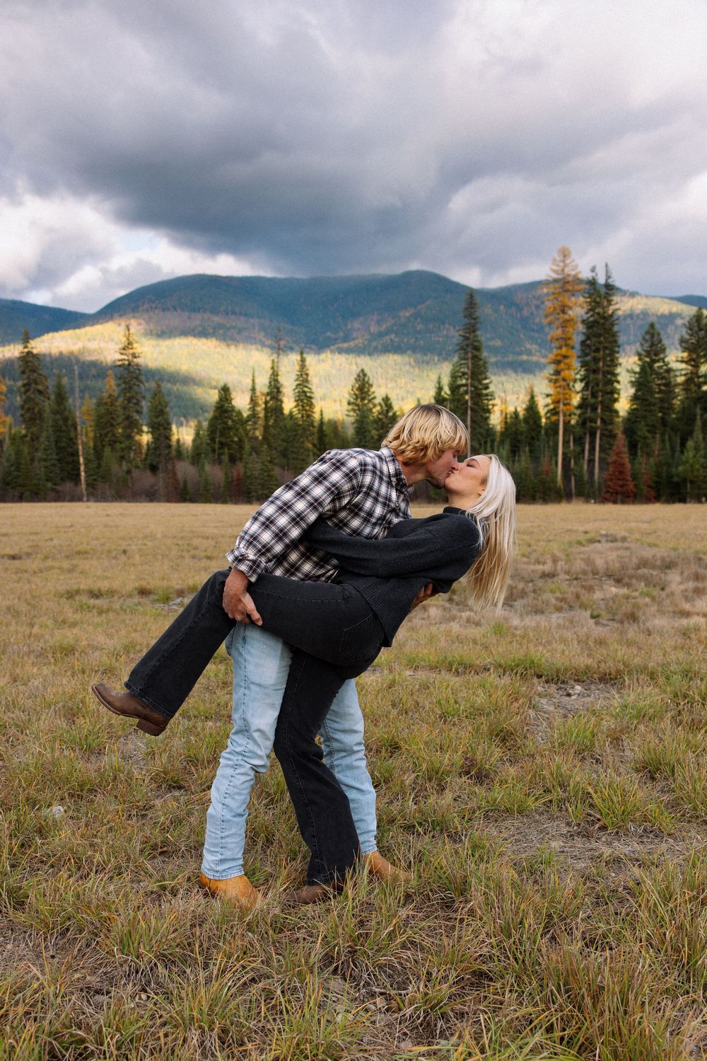 A couple embraces playfully in a mountain meadow with evergreen trees and dramatic cloudy skies in the background.