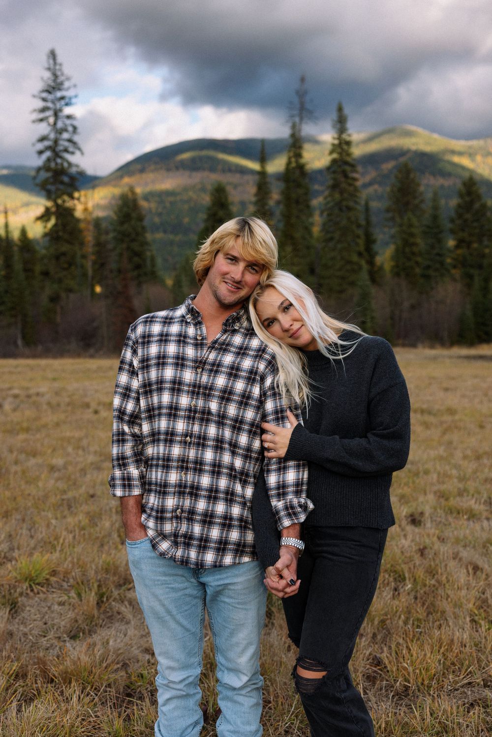 A couple stands close together in a scenic mountain meadow with evergreen trees and cloudy skies in the background.