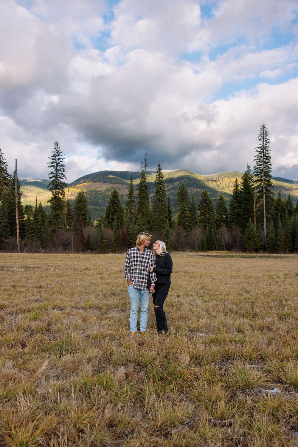 A couple stands together in a mountain meadow surrounded by evergreen trees and rolling hills under cloudy skies.