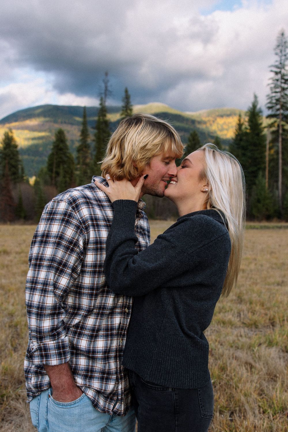 A couple shares a romantic moment in a mountain meadow with evergreen trees and moody clouds in the background.