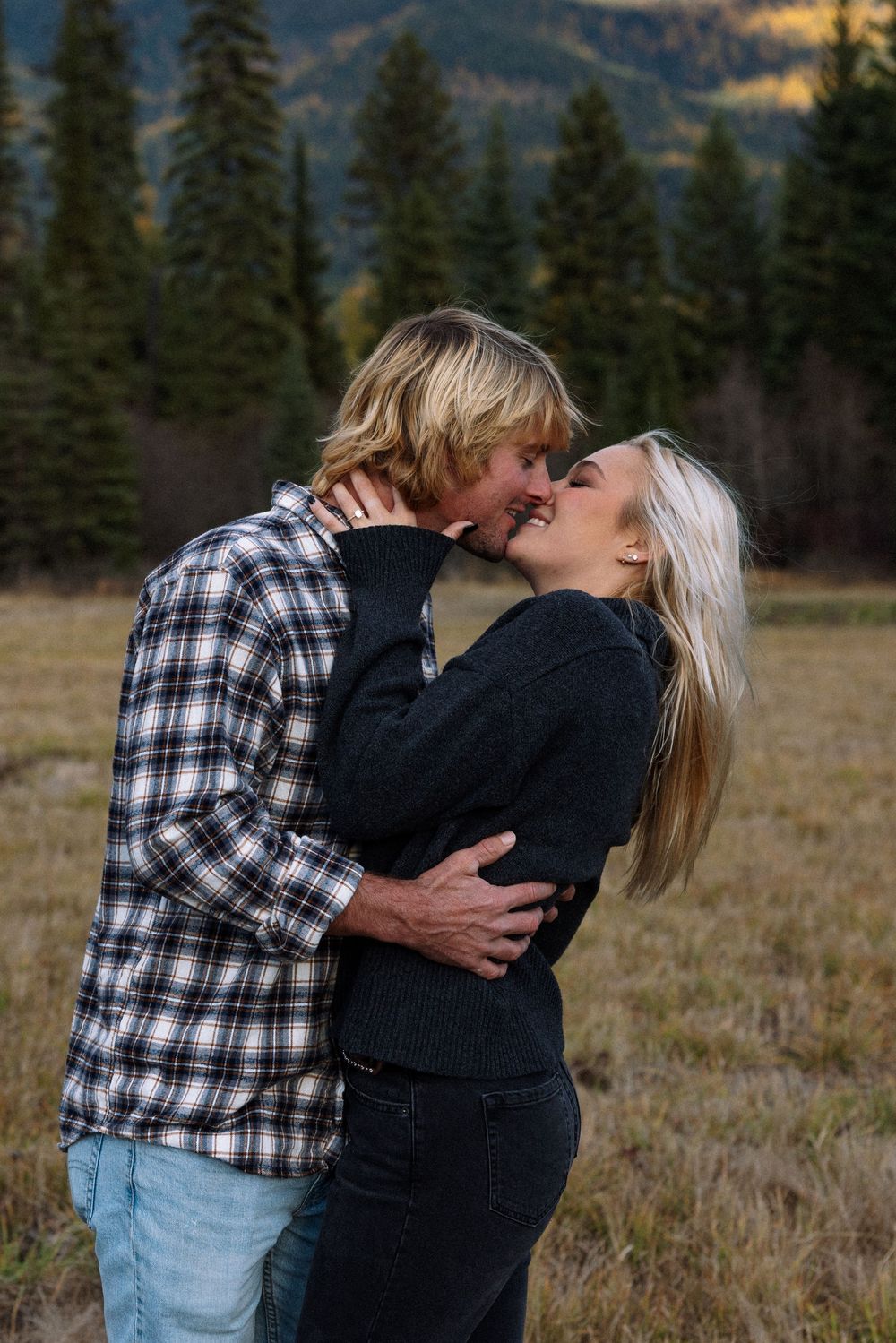 A romantic couple shares a tender kiss in a mountain meadow surrounded by evergreen trees at sunset.