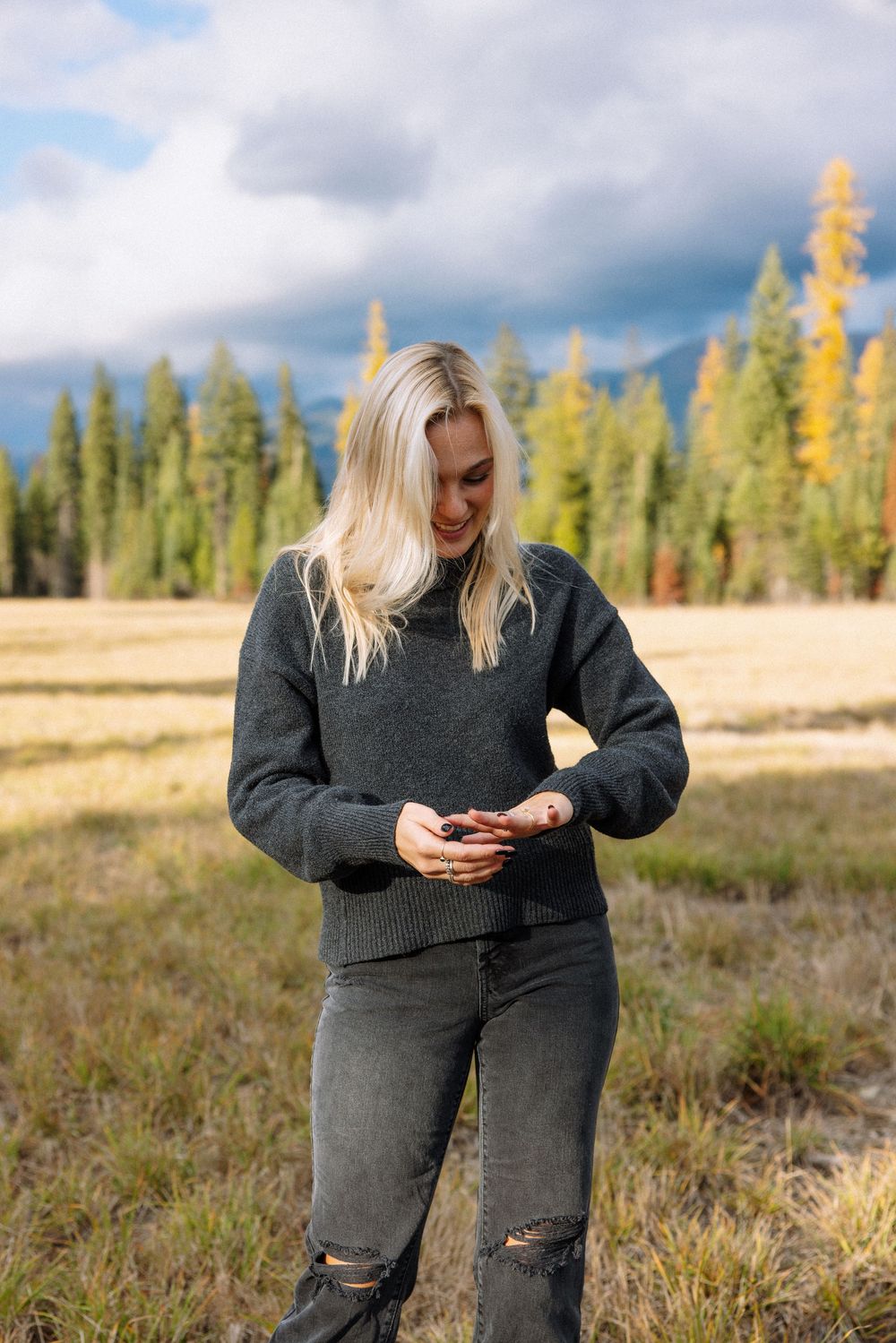 Person in gray sweater checks phone while standing in mountain meadow with evergreen forest and autumn colors.