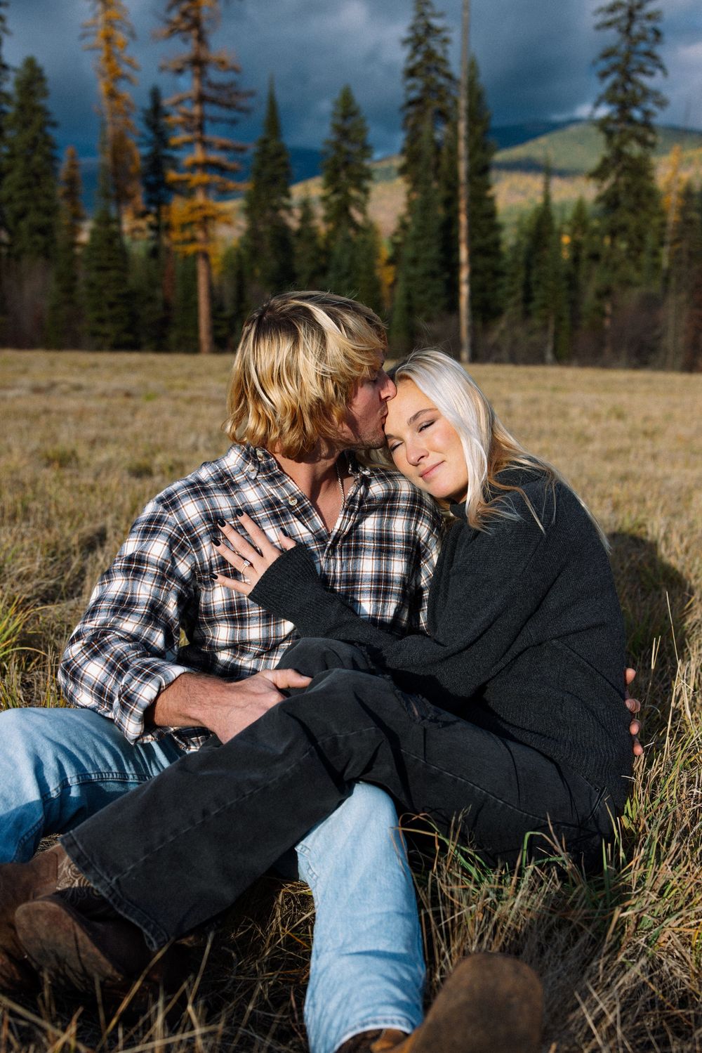 A couple shares an intimate moment while sitting in a mountain meadow with evergreen trees and stormy skies in the background.