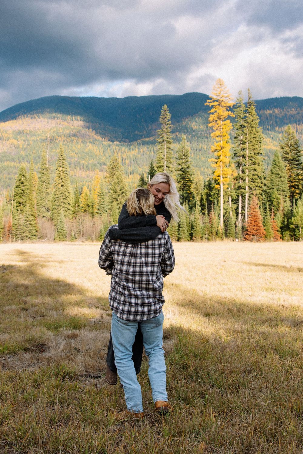 Someone in a plaid shirt and jeans stands in a meadow surrounded by evergreen trees and mountains on a cloudy day.