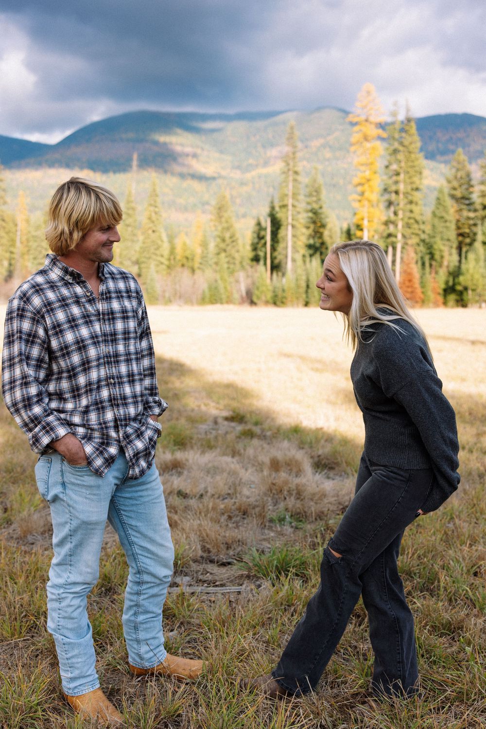 Two people in casual wear having a conversation against a backdrop of mountains and evergreen trees in autumn.