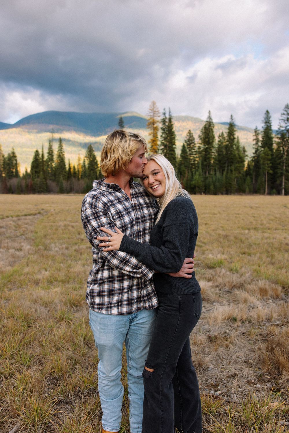 A couple embraces in a scenic meadow with mountains and evergreen trees in the background during golden hour.