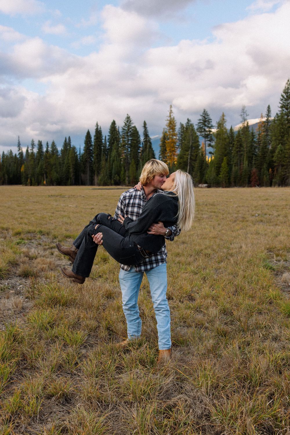 Romantic couple embracing in a meadow with evergreen trees and cloudy sky in the background.