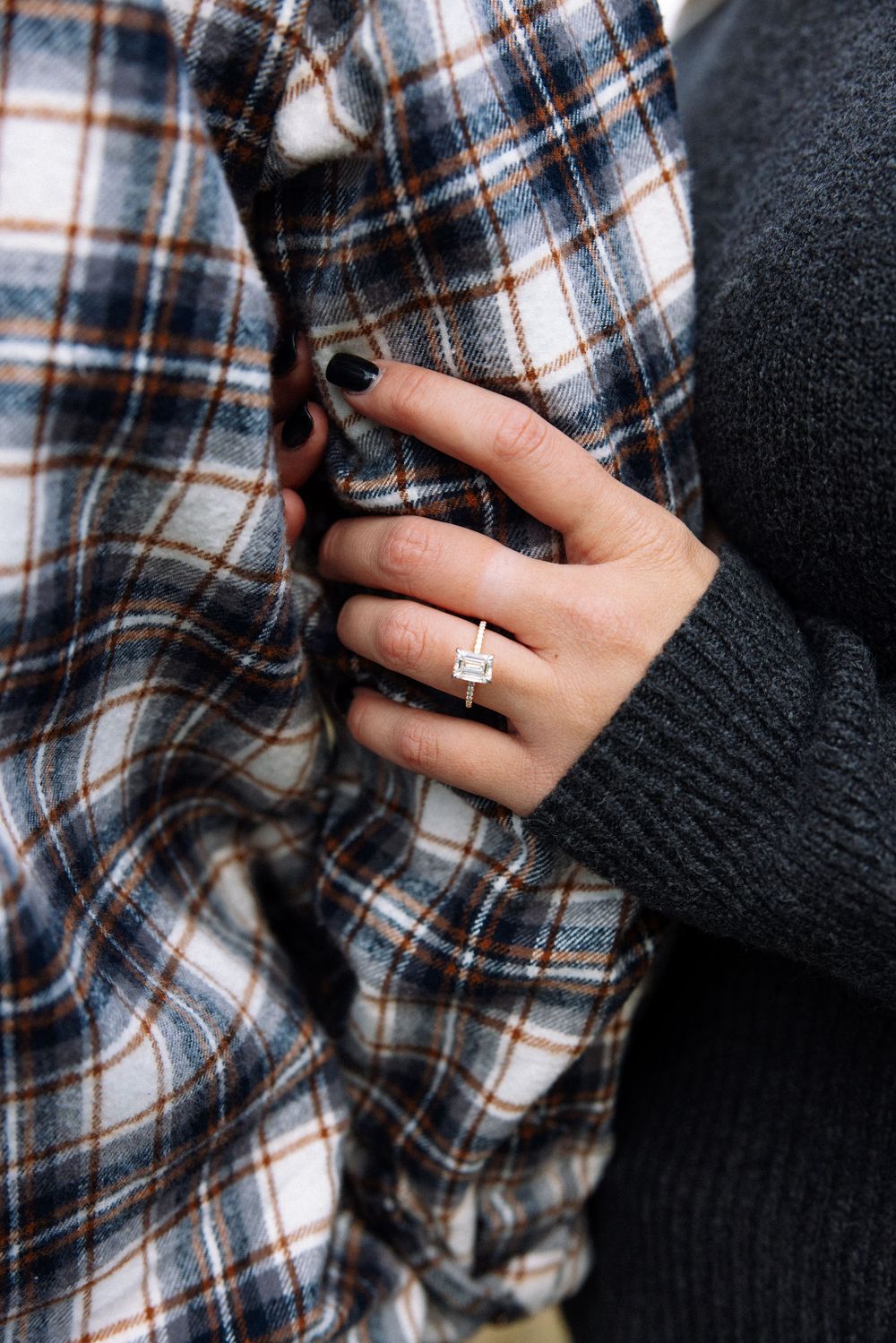 Close-up of a person wearing a plaid flannel shirt and black sweater, showing a diamond engagement ring on their hand.