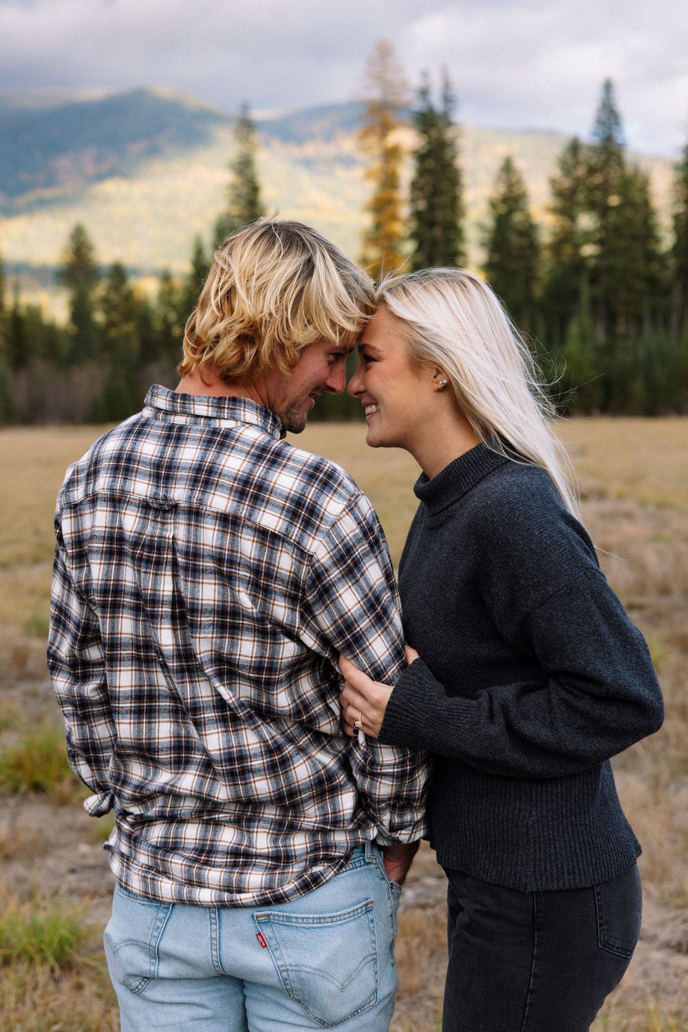 A couple in casual outdoor wear embrace intimately in a mountain meadow with evergreen trees and peaks in the background.