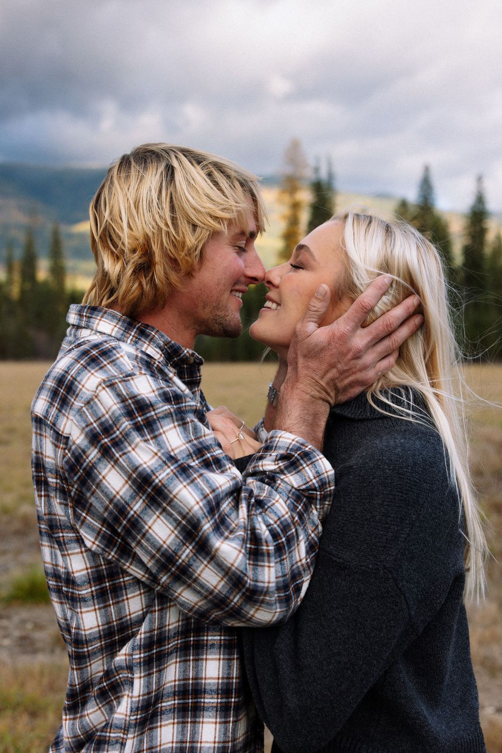 A romantic couple shares an intimate moment outdoors with mountains and evergreen trees in the background.