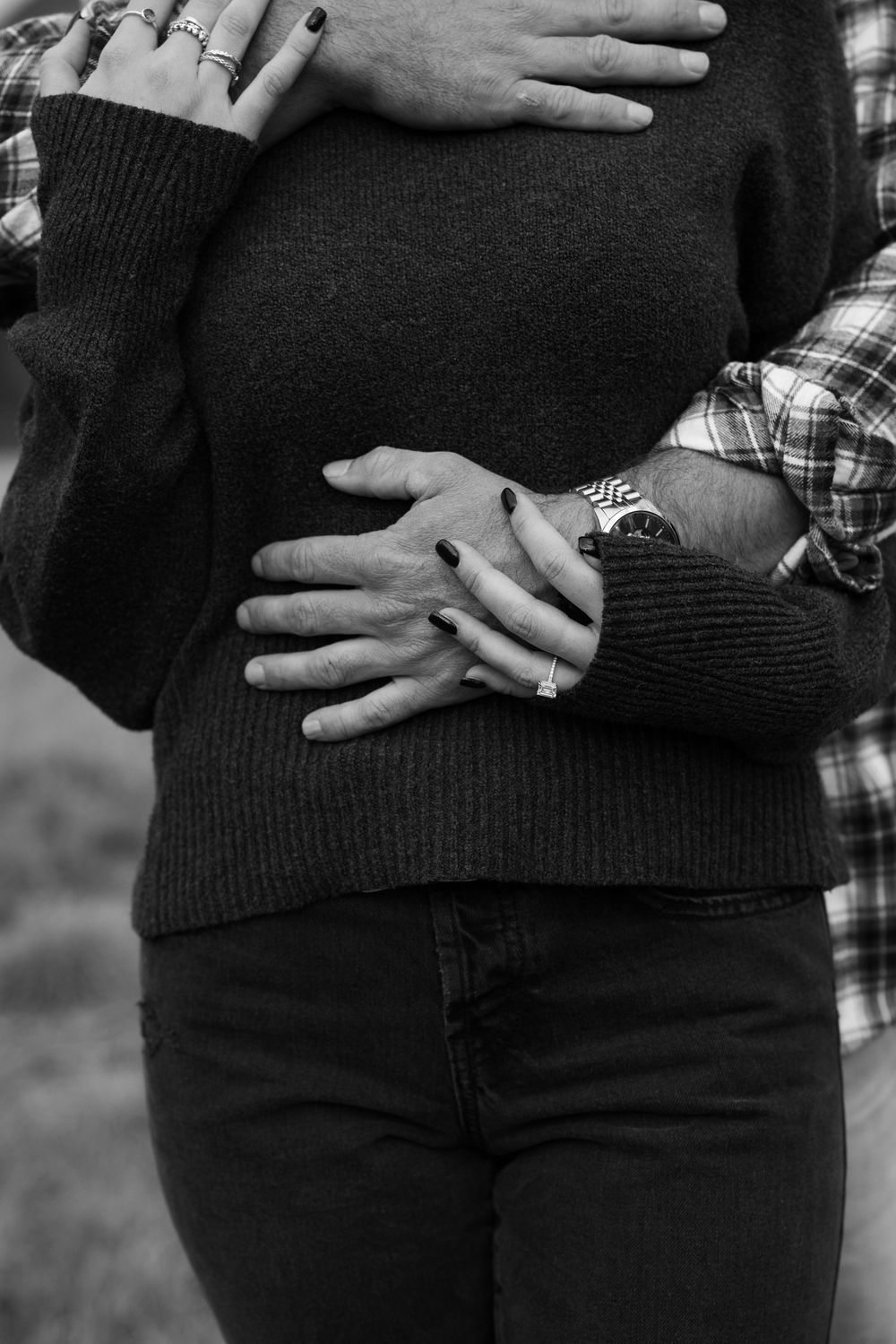 Black and white close-up shot of hands embracing a sweater-clad torso with an engagement ring sparkling on display.