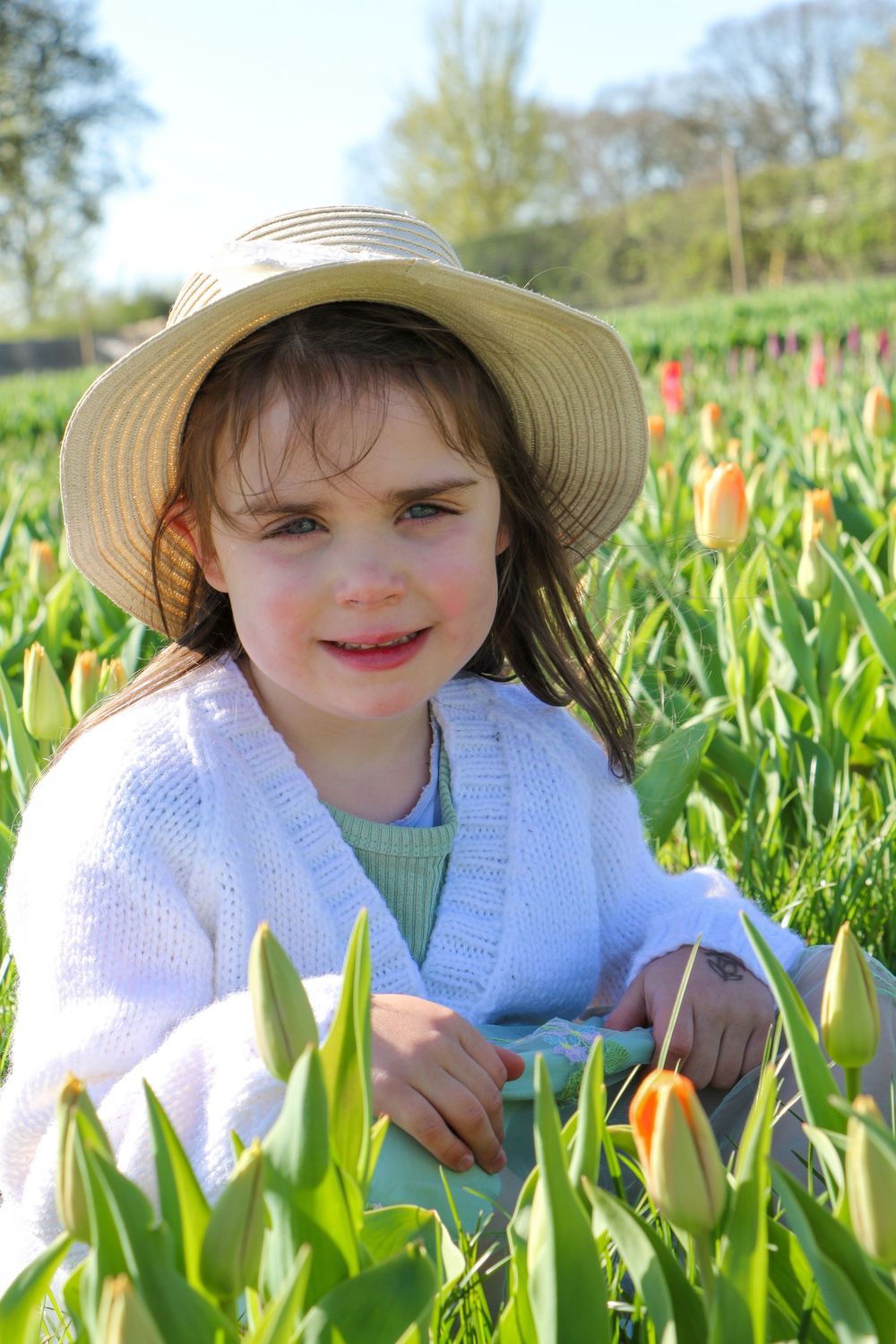 Child in a sun hat sits among colorful tulips in a springtime garden.
