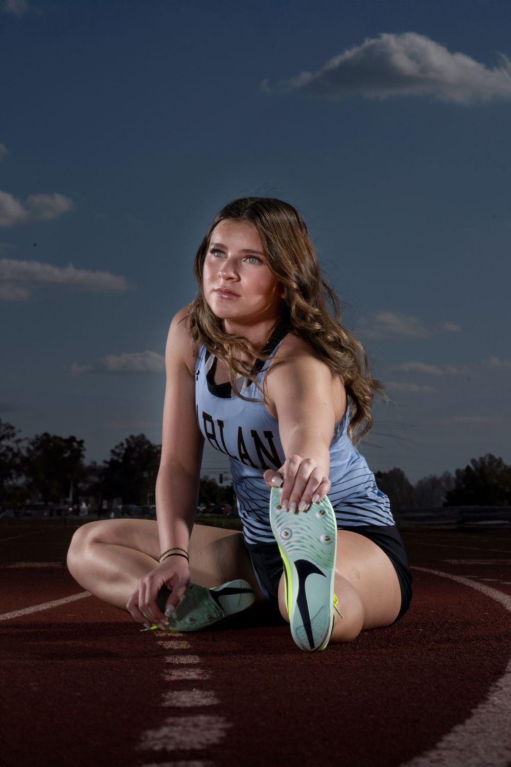 An athlete in light blue track uniform stretches on a running track at dusk with clouds in the background.