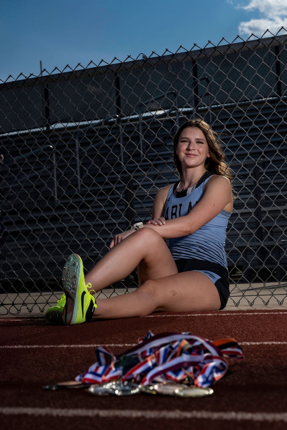 Track and field athlete rests on running track with bright neon shoes and pile of medals in dramatic lighting.