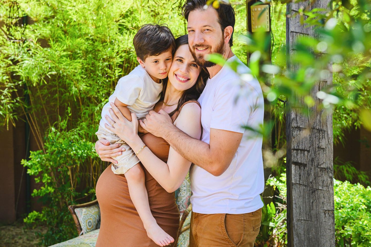 A family shares a warm embrace in a lush green garden setting with sunlight filtering through the foliage.