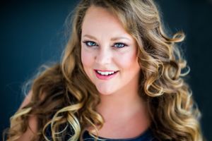 Professional headshot of person with curly blonde hair against dark blue background.
