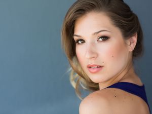 Professional headshot with soft makeup and elegant updo against blue backdrop.